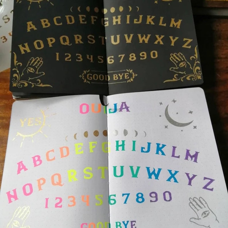 May include: Two open notebooks with a Ouija board printed on each page. The top notebook has a black background with gold lettering and symbols. The bottom notebook has a white background with colorful lettering and symbols. The word "OUIJA" is printed in rainbow colors at the top of the bottom notebook.