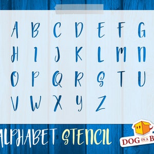 Op de afbeelding: Witte stencil met het alfabet in een handschriftlijk lettertype. De stencil is op een blauwe houten achtergrond. De tekst "ALPHABET STENCIL" staat onderaan de stencil. De tekst "DOG IN A BOX" staat rechtsonder in de afbeelding.