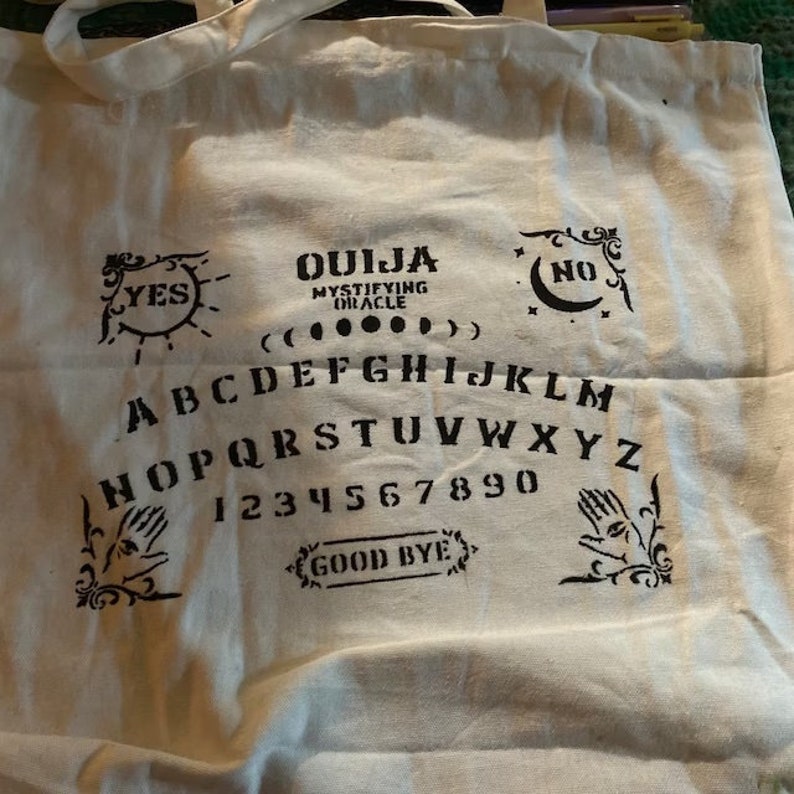 May include: A white canvas tote bag with a black Ouija board design. The design includes the letters of the alphabet, numbers, and the words "Yes", "No", and "Good Bye".
