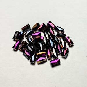 Könnte beinhalten: Ein Haufen irisierender schwarzer und violetter Glasperlen. Die Perlen sind facettiert und haben eine rechteckige Form.