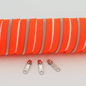 Könnte beinhalten: Nahaufnahme einer Stapel orangefarbene Reißverschlussband mit silbernen Zähnen. Drei silberne Reißverschluss-Schieber sind unterhalb des Bandes zu sehen.