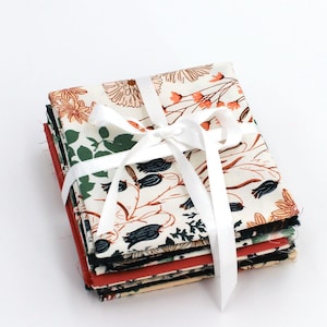 Könnte beinhalten: Ein Stapel gemusterter Stoffquadrate, gebunden mit einem weißen Band. Die Stoffe zeigen Blumen- und Pflanzenmuster in Creme, Orange, Grün und Marineblau. Der Stapel wird vor einem weißen Hintergrund präsentiert.