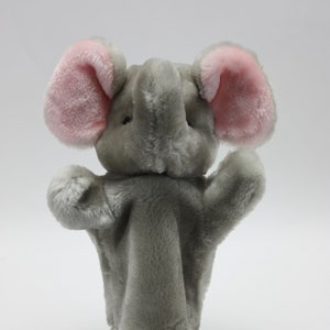 Può includere: Marionetta a mano di elefante di peluche grigia con orecchie rosa. La marionetta è in piedi su una base di ventosa trasparente.