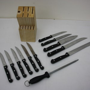 Op de afbeelding: Een set keukmessen met zwarte handvatten en een houten messenblok. De set bevat een broodmes, een koksmes en verschillende steakmessen.