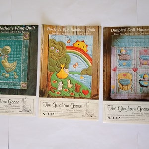 Könnte beinhalten: Drei Vintage-Quiltmusterbücher mit bunten Illustrationen. Die Cover zeigen eine Gans, einen Regenbogen und ein Puppenhaus. Die Bücher sind betitelt mit "Under Mother's Wing Quilt", "Rock-A-Bye Rainbow Quilt" und "Dimples' Doll House Quilt". Jedes Buch ist mit "The Gingham Goose Patterns & Pretties VIP" gekennzeichnet.