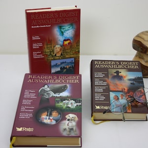 Könnte beinhalten: Drei rote Hardcover-Bücher mit goldenen Schriftzügen und Bildern auf den Umschlägen. Die Bücher tragen den Titel "Reader's Digest Auswahlbücher". Die Bücher liegen auf einer weißen Oberfläche mit einem pilzförmigen Holzobjekt und einer Brille.