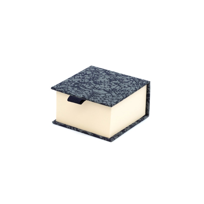 May include: A small, square box with a blue and gray patterned lid and a cream-colored interior. The box has a black ribbon pull.