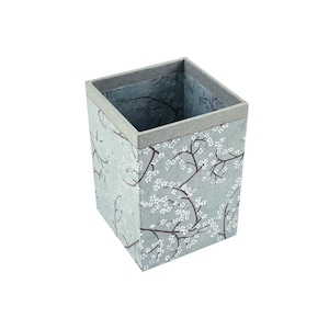 Peut inclure: Une boîte carrée en carton gris avec un motif floral. La boîte est ornée d'une bordure en papier brun autour du bord supérieur.