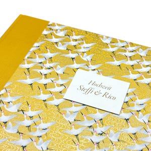 May include: A mustard yellow photo album featuring a repeating pattern of white cranes with red accents and gold wave details. A white rectangular label is attached to the front cover with the black printed text: Hochzeit Steffi & Rico.