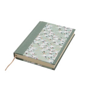 Può includere: Un diario con copertina rigida, dorso in tessuto verde salvia e una copertina decorata con un motivo ripetuto di gru bianche con dettagli rossi su sfondo azzurro con ghirigori dorati. Un sottile segnalibro a nastro beige sporge fuori.