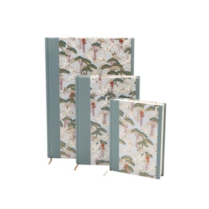 Pode incluir: Três diários de capa dura de tamanhos variados, cada um com uma lombada de tecido azul-claro e um painel central decorado com um padrão de grous brancos, pinheiros verdes e flores de glicínia roxas pendentes sobre um fundo azul-pálido.