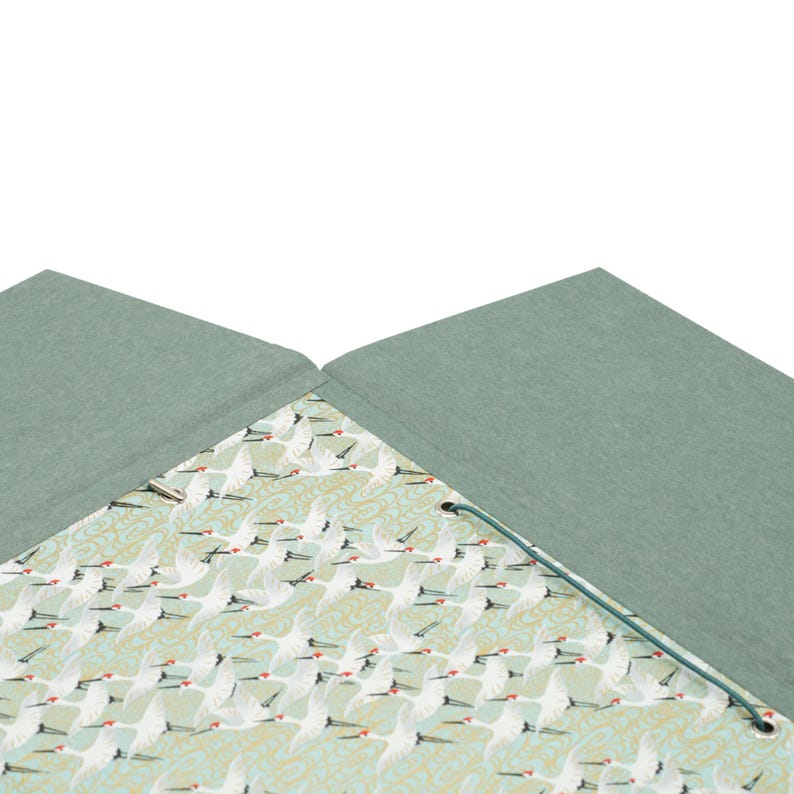 Op de afbeelding: Een saliegroene, met stof beklede notitieboek of map met een binnenvoering van een herhalend patroon van witte kraanvogels met rode accenten op een lichtgroene achtergrond, vastgezet met een dun donkergroen elastisch koord door metalen oogjes.