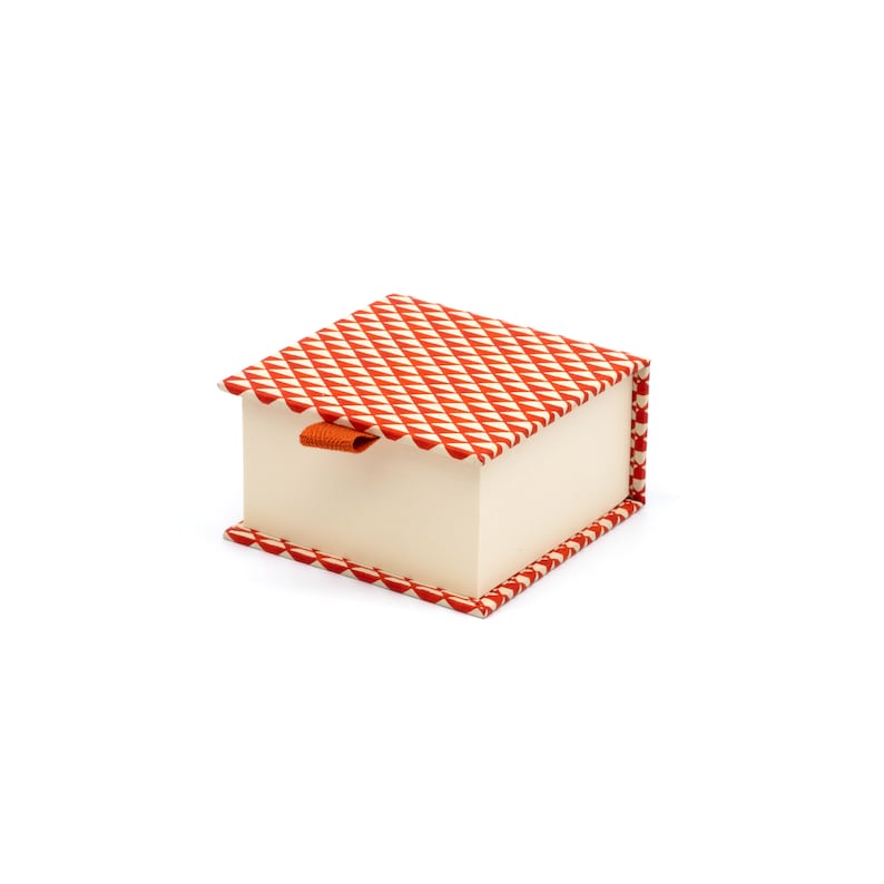 May include: A small, square box with a red and white geometric pattern. The box has a cream-coloured interior and a small orange ribbon pull.