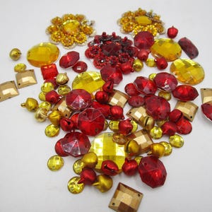 Könnte beinhalten: Eine Sammlung verschiedener Bastelverzierungen in Rot-, Gold- und Bernsteintönen. Das Sortiment umfasst facettierte Edelsteine, kleine Glöckchen und dekorative Blumenelemente, ideal für Schmuck oder andere Bastelprojekte.