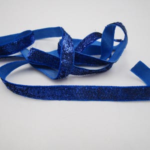 Könnte beinhalten: Ein königsblaues Band mit glitzernder Textur. Das Band ist geschlungen und auf einer weißen Oberfläche angeordnet. Das Band besteht aus einem gewebten Material und hat ein funkelndes Finish.
