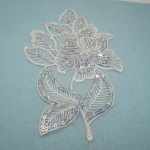 Könnte beinhalten: Weiße Spitzenblumenapplikation mit schimmernden Pailletten. Die Blume hat ein zartes Design und ist perfekt, um jedem Projekt einen Hauch von Eleganz zu verleihen.