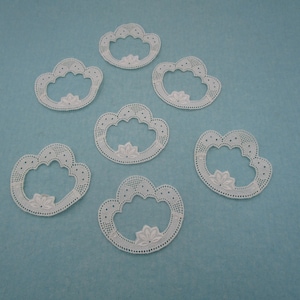 Könnte beinhalten: Sechs weiße Spitzendeckchen in Wolkenform mit einem gewellten Rand. Die Deckchen sind in einem Halbkreis auf einem hellblauen Hintergrund angeordnet.
