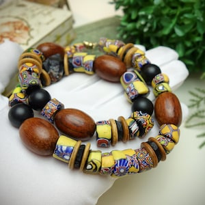 May include: A handcrafted necklace featuring a mix of wooden, black, and patterned glass beads. The necklace includes large, oval-shaped wooden beads, interspersed with smaller, colorful glass beads in yellow, blue, and red. The necklace is displayed on a white surface.