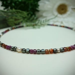 Puede incluir: Un delicado collar con una hebra de pequeñas cuentas de colores. Las cuentas incluyen tonos de morado, naranja, gris y blanco, con un solo acento de perla. El collar está diseñado para ser usado alrededor del cuello.