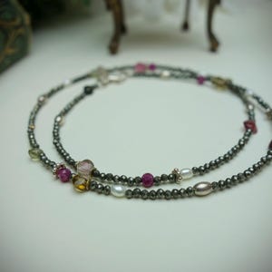 Op de afbeelding: Twee strengen kralenkettingen met een verscheidenheid aan kleurrijke edelstenen en parels. De kralen zijn donkergrijs van kleur en de edelstenen omvatten roze, gele en heldere stenen. De kettingen worden op een wit oppervlak getoond.