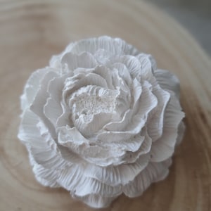 May include: A white, ceramic flower with a ruffled texture. The flower is on a light brown wooden surface.