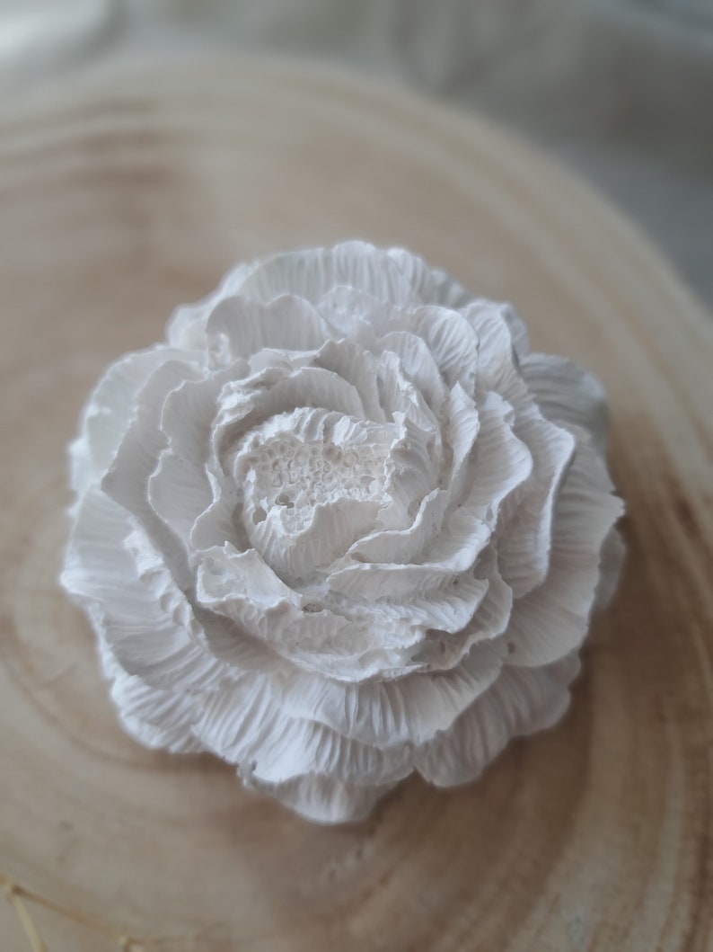 May include: A white, ceramic flower with a ruffled texture. The flower is on a light brown wooden surface.