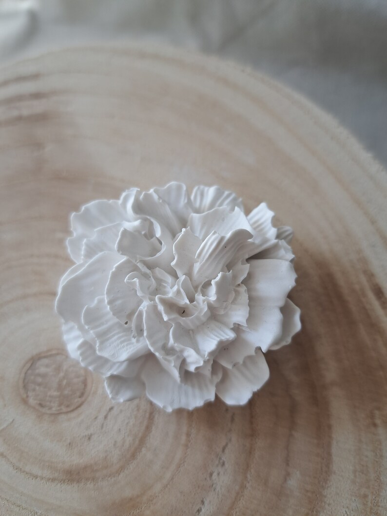 May include: A white ceramic flower with a ruffled texture. The flower is on a light brown wooden surface.
