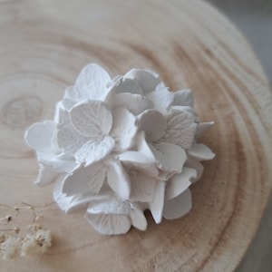 May include: A white ceramic hydrangea flower, with a detailed design, sits on a light brown wooden surface.