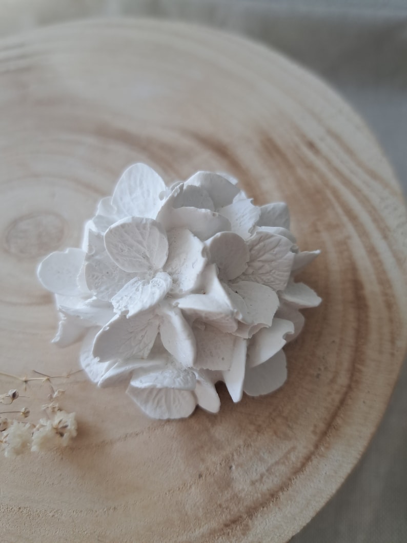 May include: A white ceramic hydrangea flower, with a detailed design, sits on a light brown wooden surface.