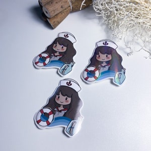 Puede incluir: Tres pegatinas iridiscentes con una niña de dibujos animados que lleva un gorro de marinero y sostiene un salvavidas. La niña está de pie frente a una ola azul con un mensaje en una botella.