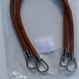 Op de afbeelding: Twee bruine leren tashendels met zilveren metalen sluitingen. De handvatten zijn gevlochten en verpakt in een doorzichtige plastic zak. De tas is gelabeld met "Manici per Borse Bag Handles".