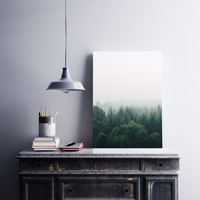 K&ouml;nnte beinhalten: Eine graue Pendelleuchte h&auml;ngt &uuml;ber einer dunklen Holzkonsole mit einem Stapel B&uuml;cher und einem Stiftehalter. Ein gerahmtes Bild einer nebligen Waldszene h&auml;ngt an der Wand hinter der Konsole.