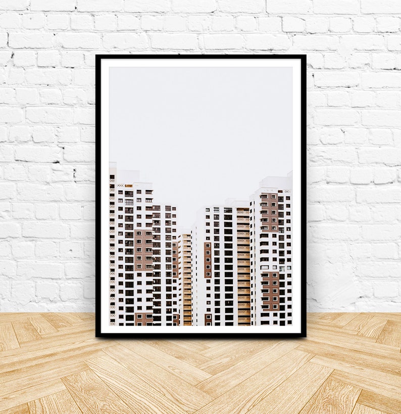 May include: Framed art print featuring a minimalist architectural photograph. The image showcases several high-rise apartment buildings with white and brown facades against a pale sky. The buildings have many windows and balconies. The print is in a black frame.