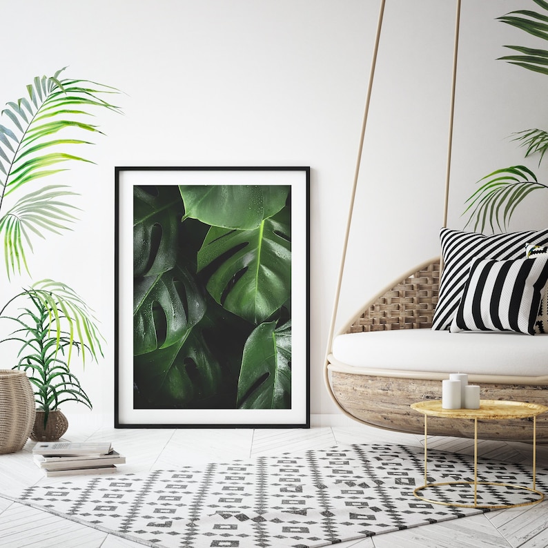 May include: A black framed print of a close-up of lush green tropical leaves. The print is hanging on a white wall above a white wicker hanging chair with black and white striped pillows.