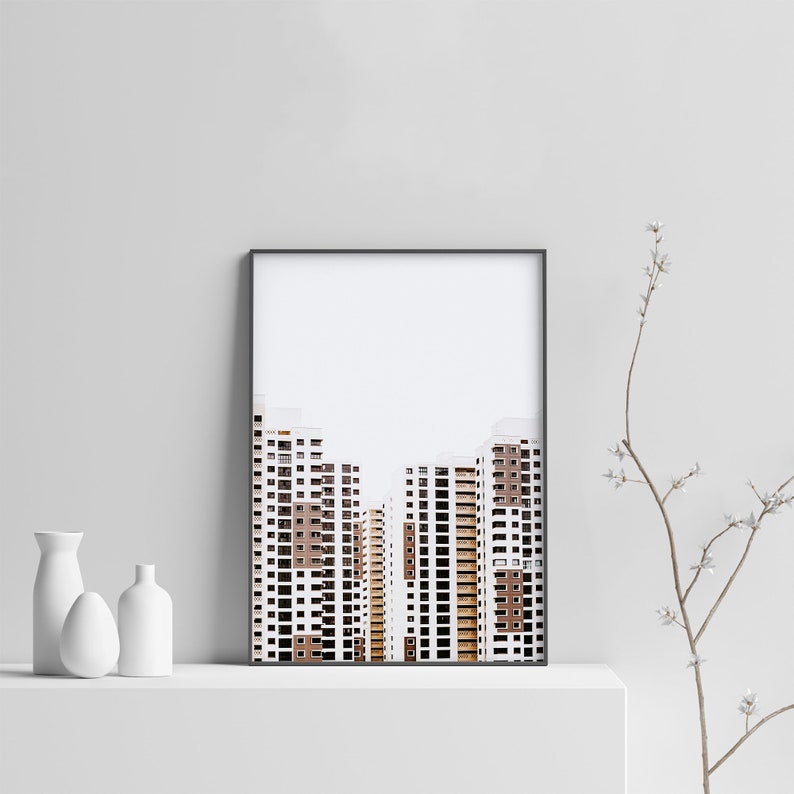 May include: Framed art print featuring a minimalist architectural photograph of several high-rise buildings. The buildings have white and brown facades with numerous windows. The print is in a black frame and is displayed on a white shelf.
