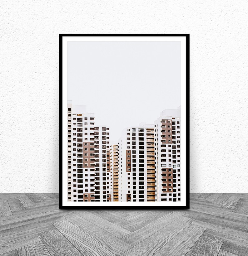 May include: Framed print featuring a minimalist architectural photograph. The image showcases several high-rise apartment buildings with white and brown facades, and numerous windows. The buildings are set against a plain, light-colored sky. The print is in a black frame.