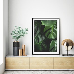 May include: A framed print of a close-up of large, dark green tropical leaves with a black frame. The print is sitting on a light wood shelf with two gold and black vases and a gold lamp.