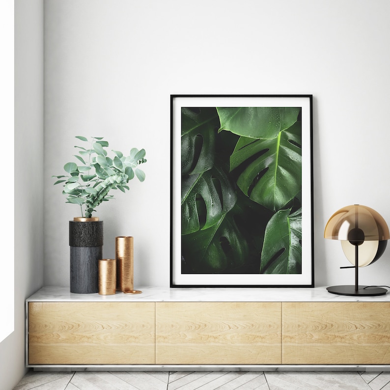 May include: A framed print of a close-up of large, dark green tropical leaves with a black frame. The print is sitting on a light wood shelf with two gold and black vases and a gold lamp.