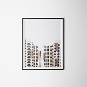 May include: Framed print featuring a photograph of several high-rise apartment buildings. The buildings are white with brown and tan accents, and have many windows. The sky is a pale gray. The frame is black.