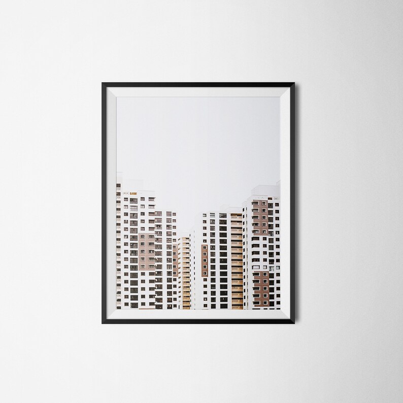 May include: Framed print featuring a photograph of several high-rise apartment buildings. The buildings are white with brown and tan accents, and have many windows. The sky is a pale gray. The frame is black.
