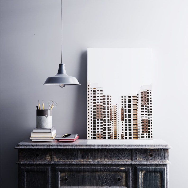 May include: A modern interior scene featuring a framed architectural print of high-rise buildings. A gray pendant light hangs above a dark wooden cabinet. On the cabinet are books, a pen, scissors, and a pencil holder.