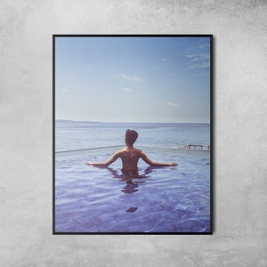 May include: A person relaxes in a swimming pool overlooking the ocean. The water is a vibrant blue, and the sky is a clear light blue. The person is facing away from the camera, looking out at the horizon.