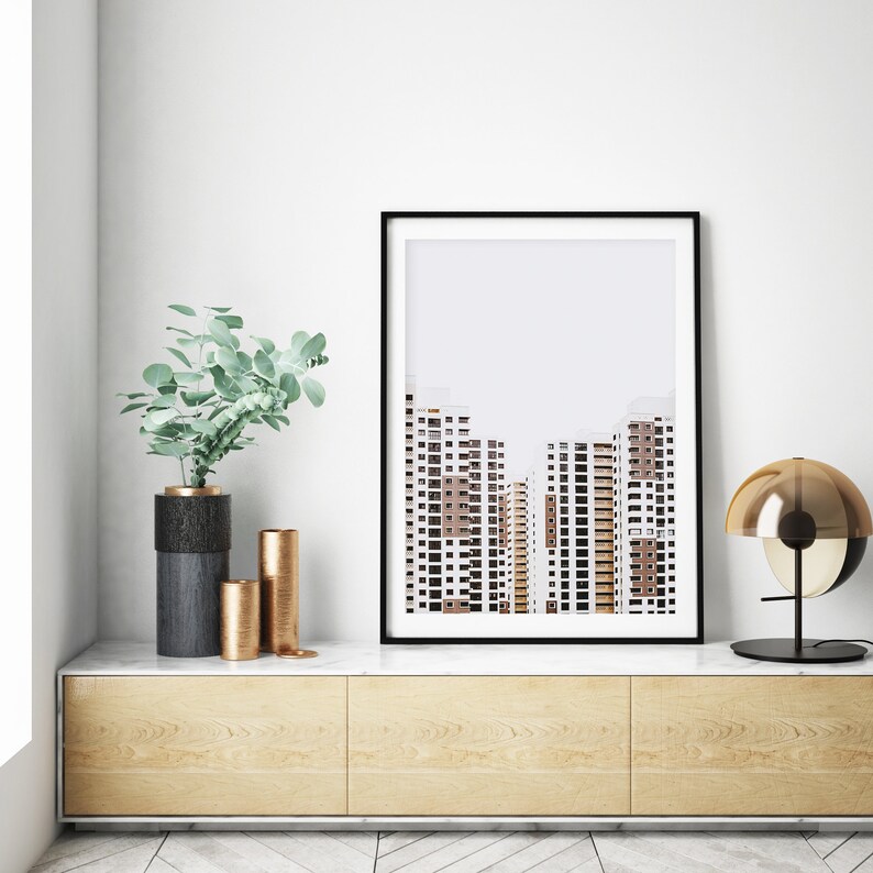May include: A framed art print featuring a minimalist architectural photograph of high-rise buildings. The print is displayed above a wooden cabinet with decorative vases and a modern lamp. The color palette includes white, brown, and black.