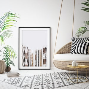 May include: A framed art print featuring a minimalist architectural design of several high-rise buildings in shades of brown and white. The print is displayed in a black frame, set against a white wall, with a patterned rug and hanging chair.