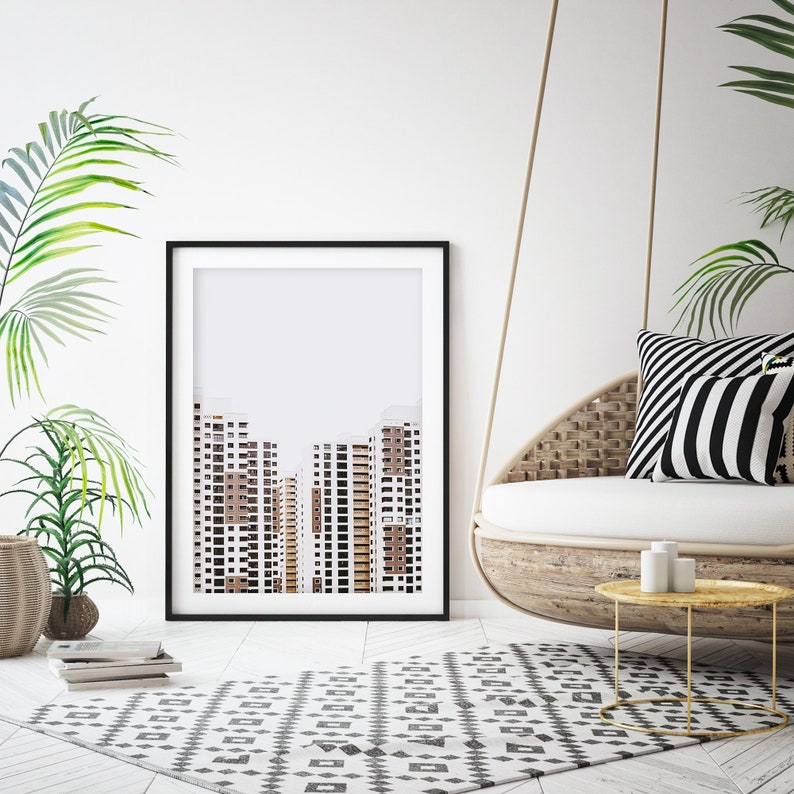 May include: A framed art print featuring a minimalist architectural design of several high-rise buildings in shades of brown and white. The print is displayed in a black frame, set against a white wall, with a patterned rug and hanging chair.