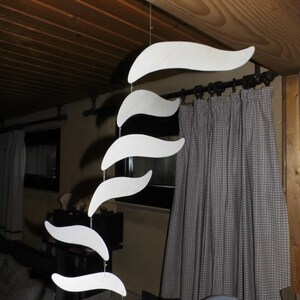 Puede incluir: M&oacute;vil de madera blanca con una serie de formas curvas, similares a olas, suspendidas de un hilo. El m&oacute;vil cuelga de un techo de madera, con una cortina de tonos neutros en el fondo. Las formas var&iacute;an en tama&ntilde;o, creando una pieza decorativa.