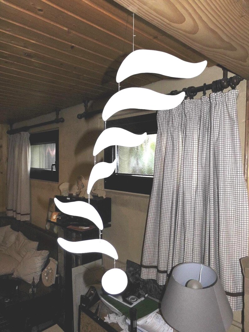 Puede incluir: Un m&oacute;vil colgante blanco moderno con una serie de formas curvas en forma de hoja suspendidas de un alambre fino. Una esfera blanca en la parte inferior. El m&oacute;vil cuelga frente a una ventana con cortinas a cuadros.
