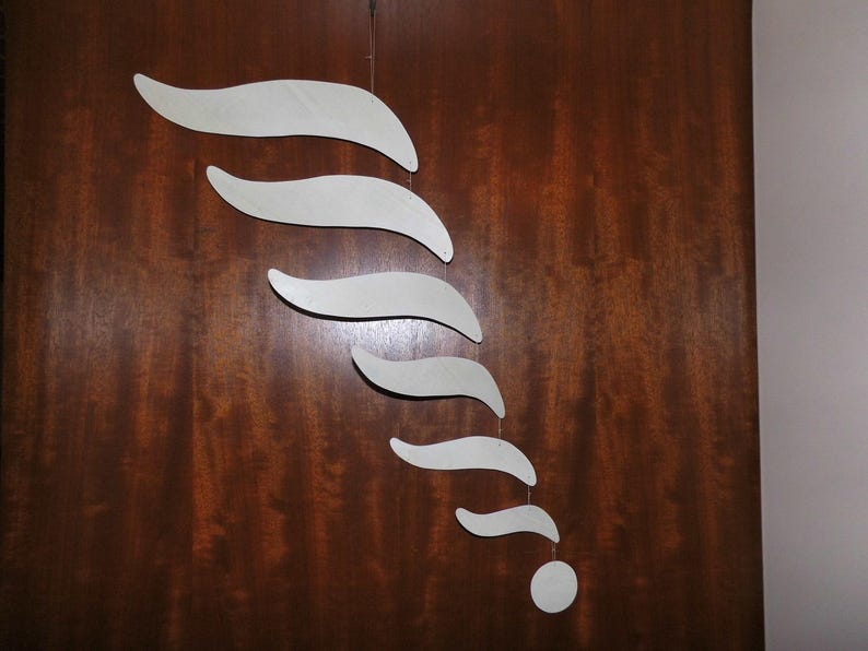 Puede incluir: M&oacute;vil abstracto con una serie de formas blancas y curvas suspendidas de un solo punto. Las formas disminuyen de tama&ntilde;o, culminando en un peque&ntilde;o disco redondo. El m&oacute;vil cuelga sobre un fondo de madera marr&oacute;n oscuro.