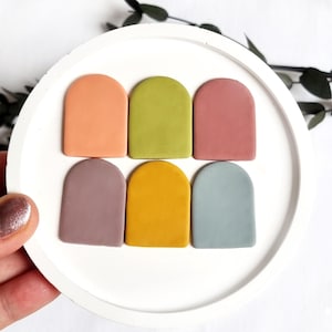 Puede incluir: Seis pequeñas formas de arcilla arqueadas en varios colores, incluyendo melocotón, verde, rosa, gris, amarillo y azul. Las formas están dispuestas en dos filas de tres sobre una superficie blanca.