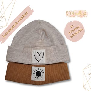 Könnte beinhalten: Eine beige Mütze mit einem braunen Umschlagrand. Die Mütze hat einen weißen Herz-Patch und einen weißen Sonnen-Patch. Der Text "Leinenpatch wählbar" ist auf einem rosa Hintergrund. Der Text "In 11 Farben erhältlich" ist auf einem rosa Hintergrund.